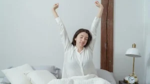 Mulher acordando na cama com expressão serena e descansada, braços levantados e luz natural suave em ambiente acolhedor.