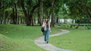 Mulher caminhando por um parque arborizado enquanto olha para o celular, em ambiente externo com luz natural suave.