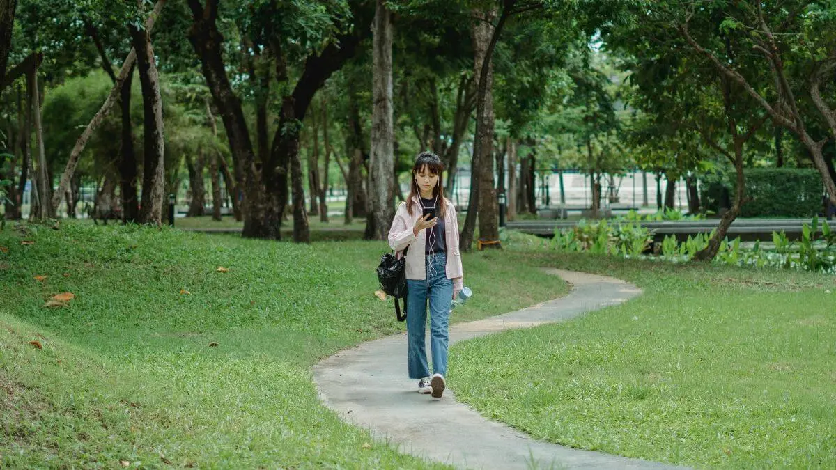 Mulher caminhando por um parque arborizado enquanto olha para o celular, em ambiente externo com luz natural suave.