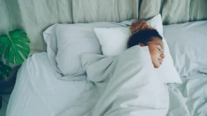 Pessoa de cabelos cacheados dormindo tranquilamente enrolada em roupa de cama branca e suave representando como dormir melhor naturalmente