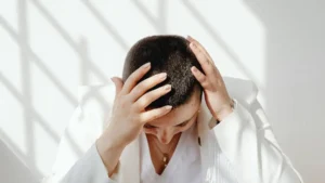Pessoa com as mãos na cabeça e olhar para baixo com luz natural suave expressando corpo cansado e mente sobrecarregada
