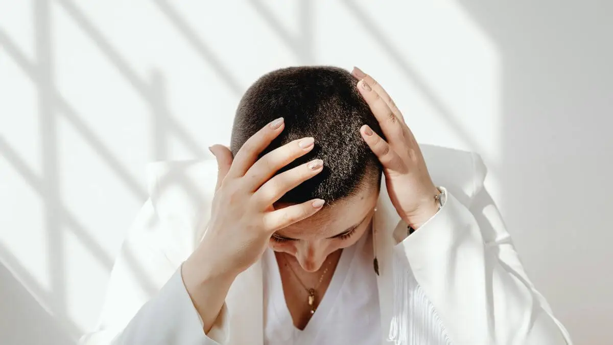 Pessoa com as mãos na cabeça e olhar para baixo com luz natural suave expressando corpo cansado e mente sobrecarregada