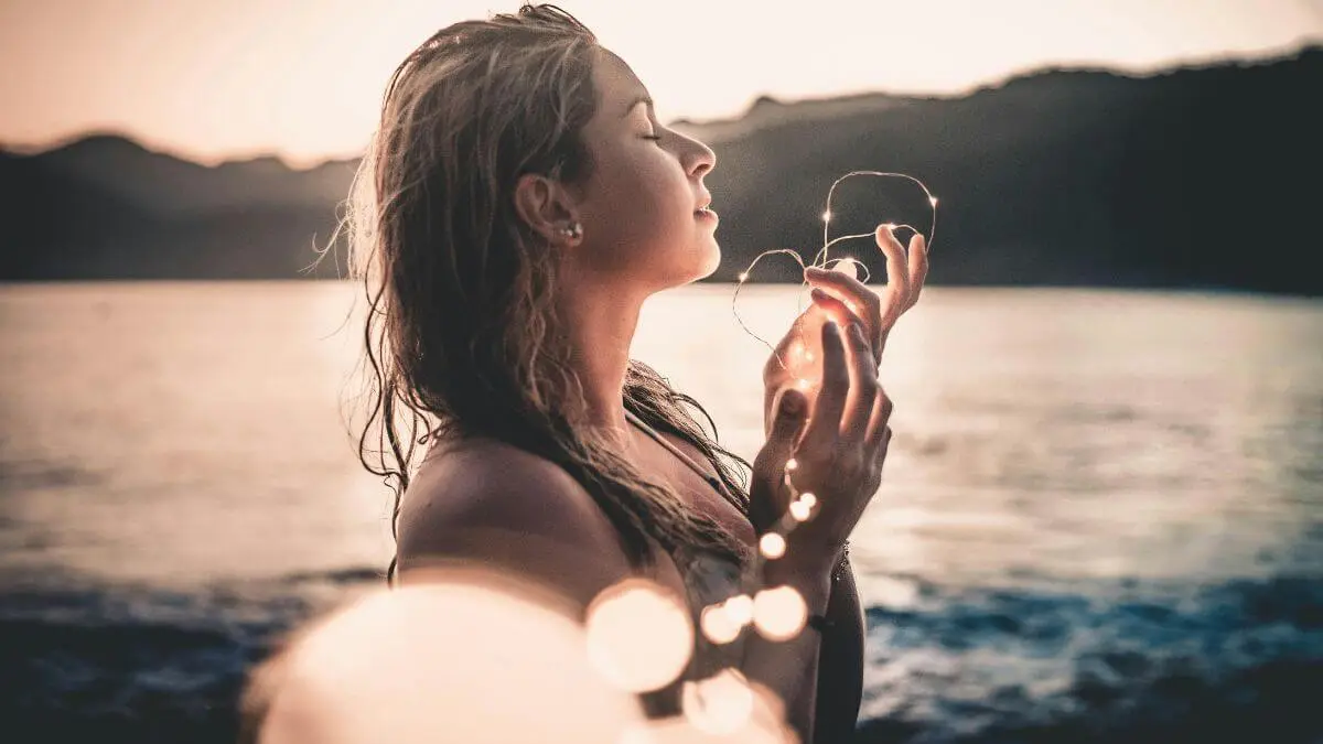 Mulher de olhos fechados com expressão serena segurando luzes ao entardecer em momento de mindfulness e presença