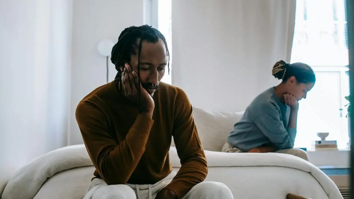 Casal sentado em lados opostos do sofá, com expressões pensativas e distantes, transmitindo distância emocional e momento difícil no relacionamento.