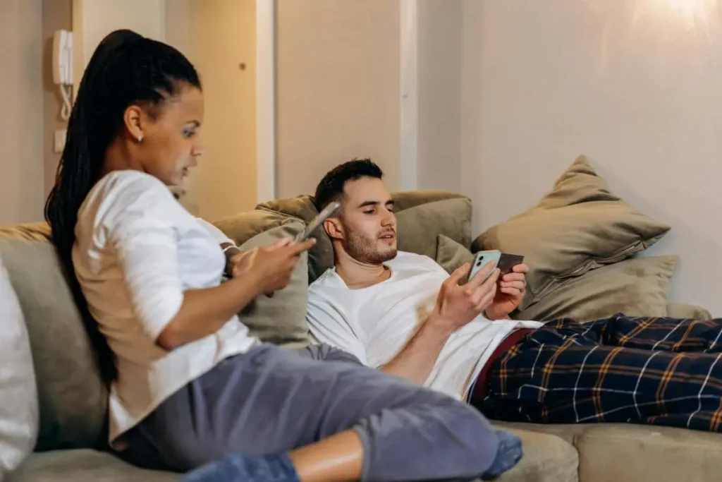 Casal em ambiente doméstico olhando para seus próprios celulares, transmitindo distância emocional e desconexão apesar da presença física.