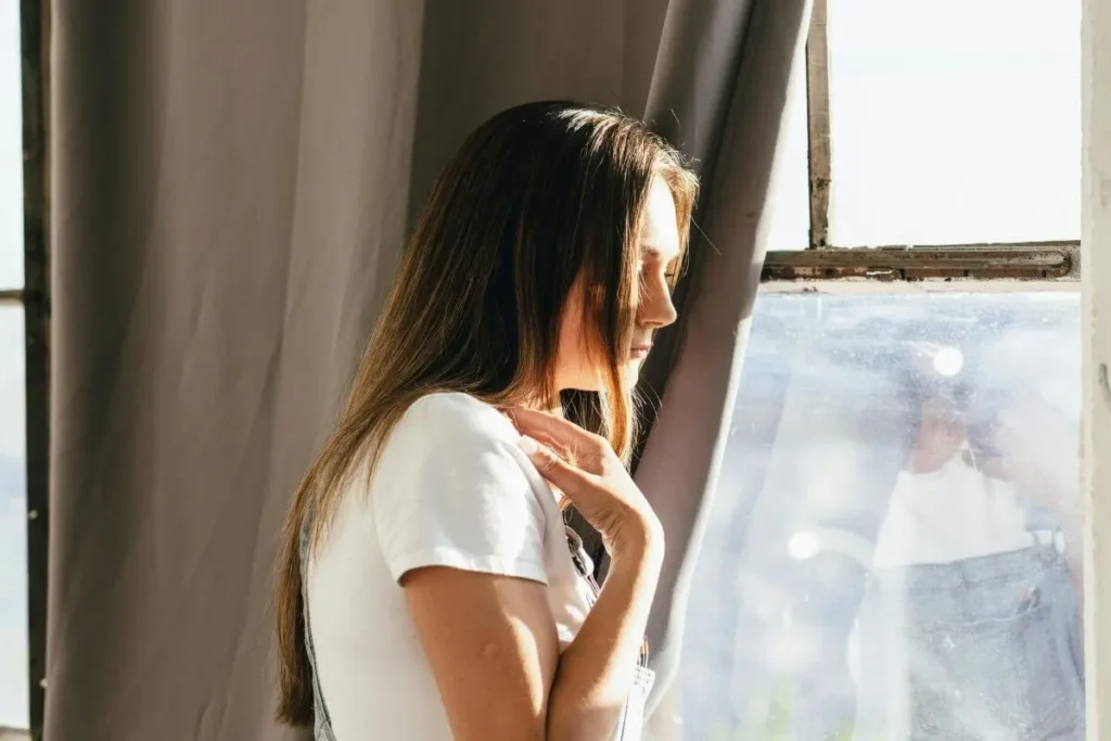 Mulher de cabelos longos olhando pela janela com expressão pensativa e cansada com luz natural suave representando sobrecarga mental e emocional