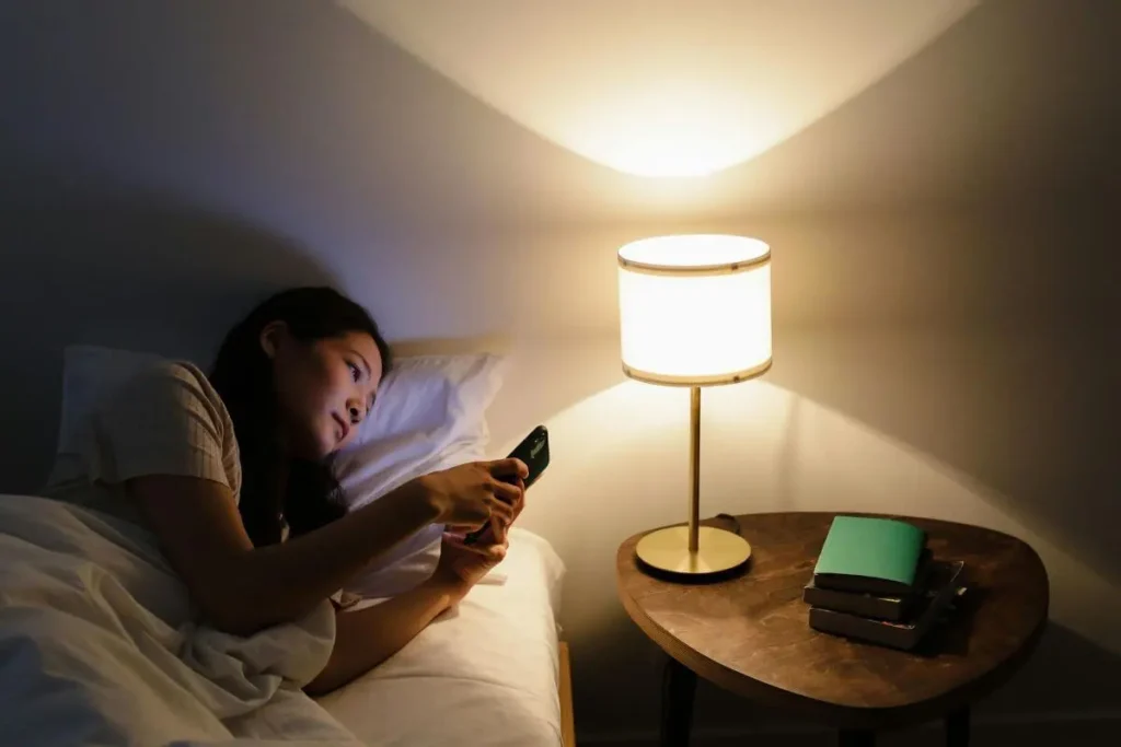 Mulher deitada na cama acordada à noite, olhando para o celular com expressão cansada, ao lado de um abajur com luz suave e quente.
