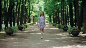 Mulher caminhando sozinha em parque arborizado, com expressão tranquila e presente, cercada por árvores e luz natural suave.