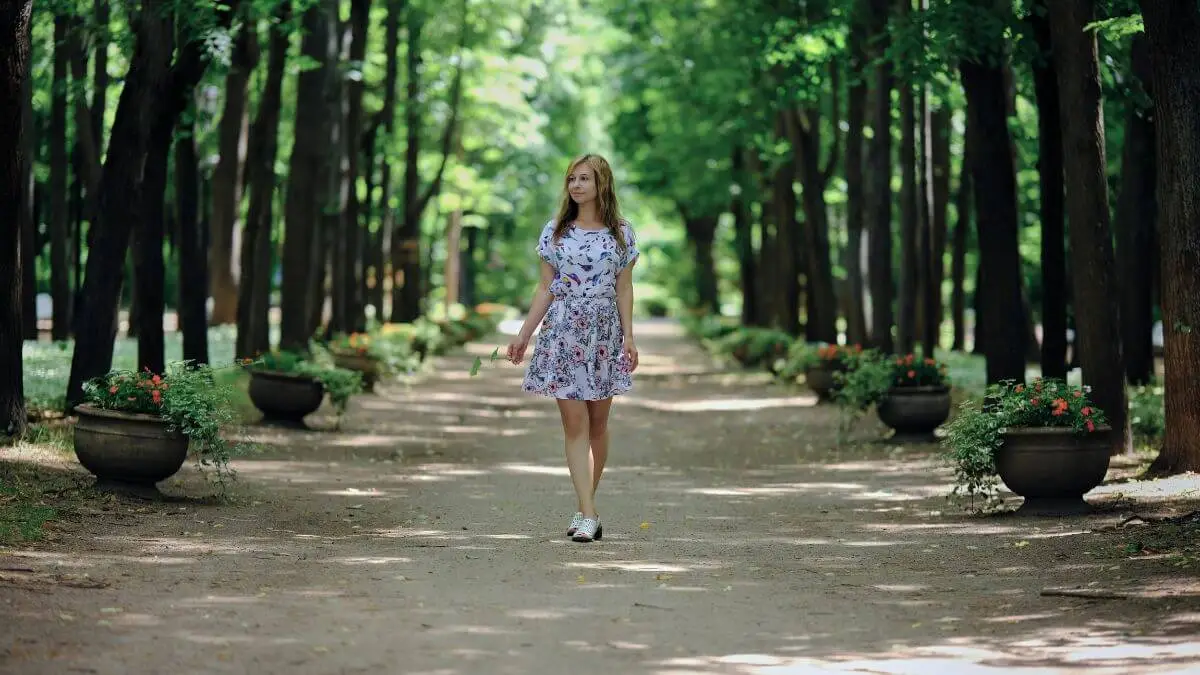 Mulher caminhando sozinha em parque arborizado, com expressão tranquila e presente, cercada por árvores e luz natural suave.