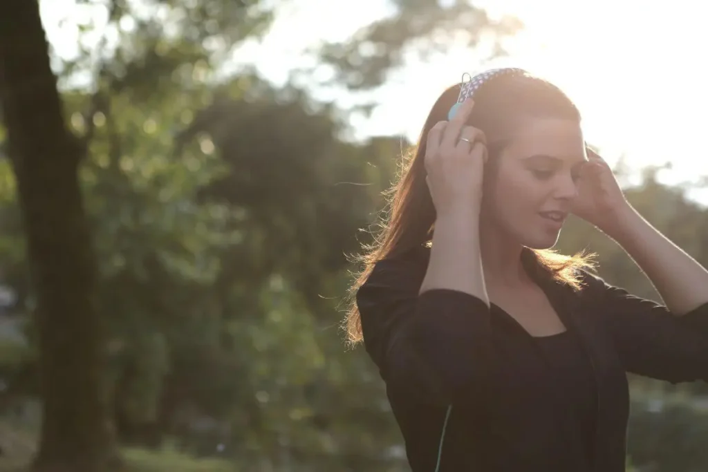 Mulher em parque usando fones de ouvido, com expressão tranquila e luz suave do fim do dia ao fundo.