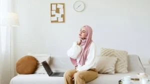 Mulher sentada no sofá com a mão no pescoço, demonstrando tensão física e cansaço em ambiente doméstico com luz suave.