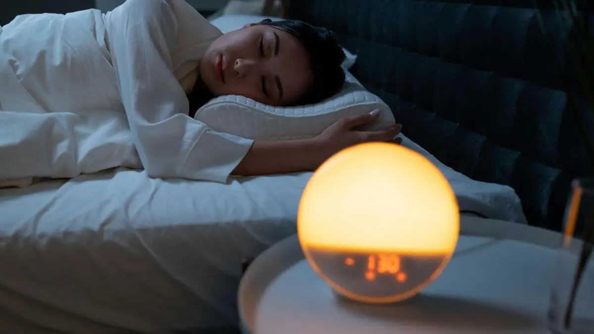 Mulher deitada na cama com olhos fechados, expressão serena e luz suave de abajur ao fundo em ambiente acolhedor.