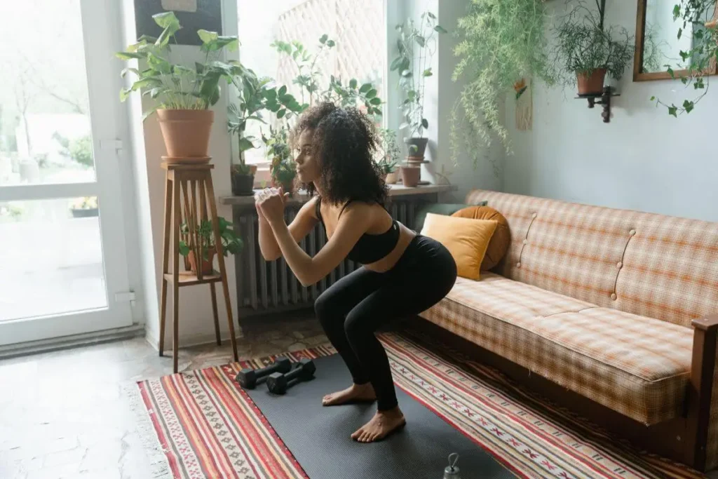 Mulher fazendo agachamento em sala de estar sobre tapete de exercícios, com postura concentrada, luz natural e plantas ao fundo.