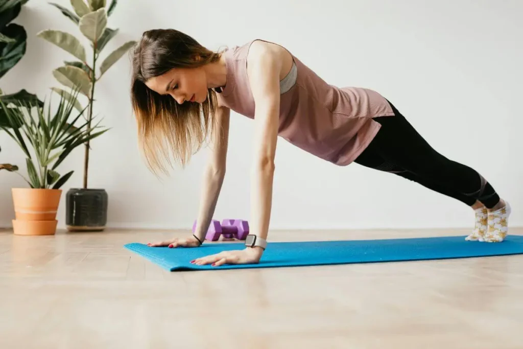 Mulher em posição de prancha sobre tapete de yoga em ambiente doméstico organizado, com expressão concentrada e planta ao fundo.