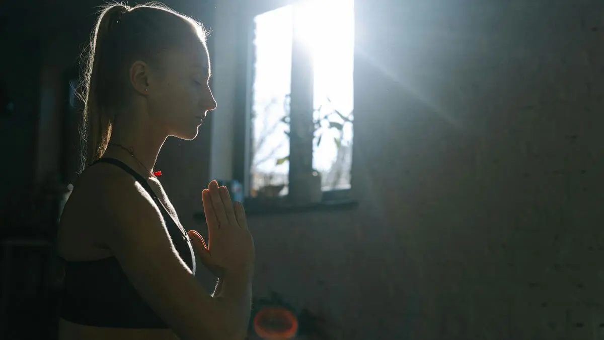 Mulher com olhos fechados e mãos unidas próximas ao peito, em momento de respiração consciente com luz natural suave ao fundo.