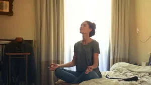 Mulher meditando no quarto com olhos fechados, sentada em posição confortável diante de uma janela com luz natural suave.