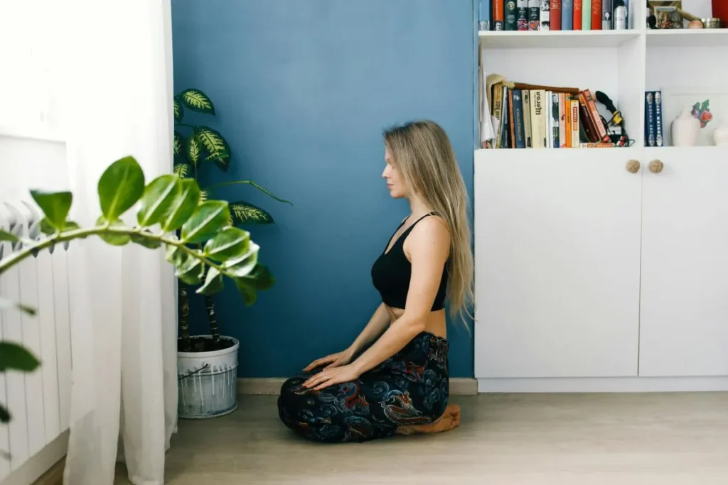 Mulher sentada no chão em posição de meditação, com as mãos sobre os joelhos, em ambiente claro com plantas e luz natural suave.
