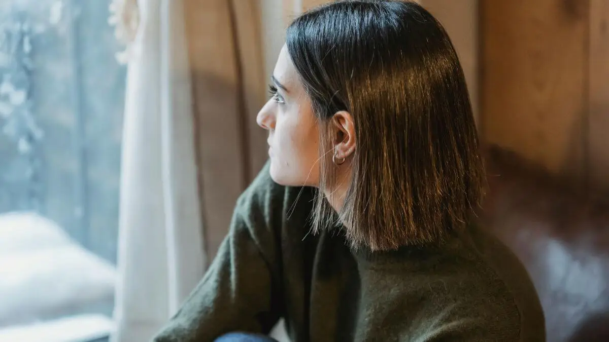 Mulher sentada próxima à janela, com olhar pensativo voltado para fora, em ambiente acolhedor com luz natural suave.