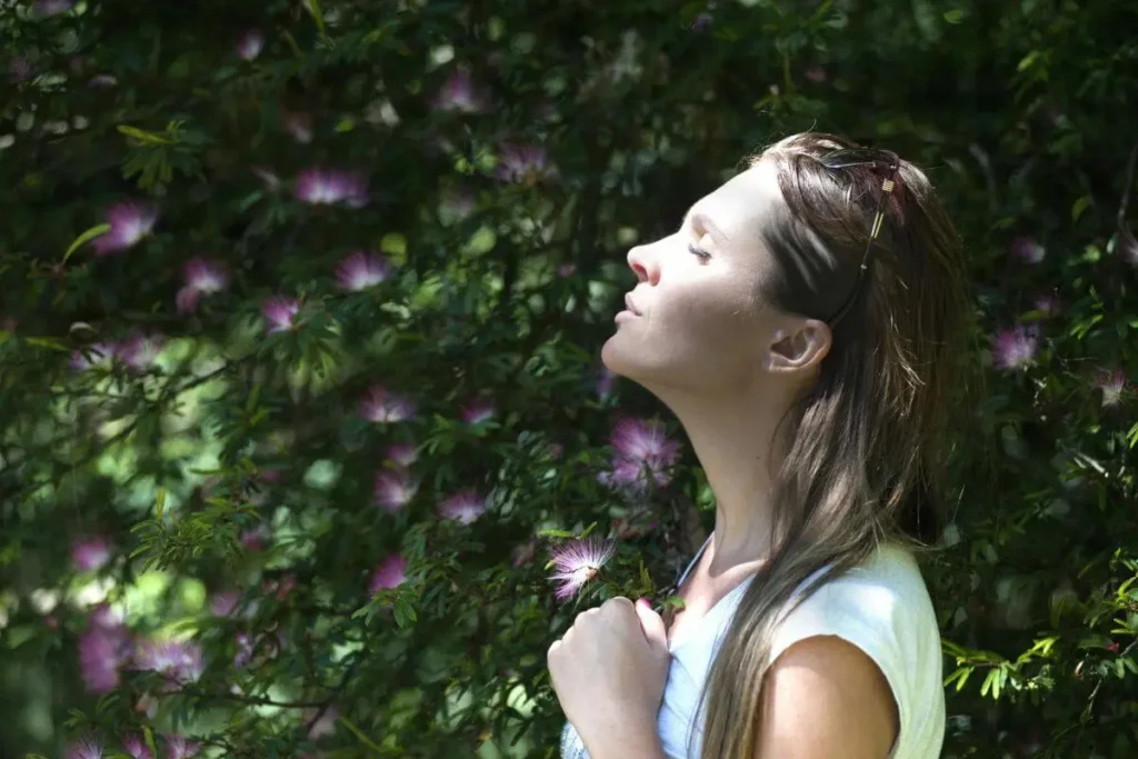 Mulher em parque com olhos fechados e expressão serena, cercada por vegetação, transmitindo presença, calma e conexão com a natureza.