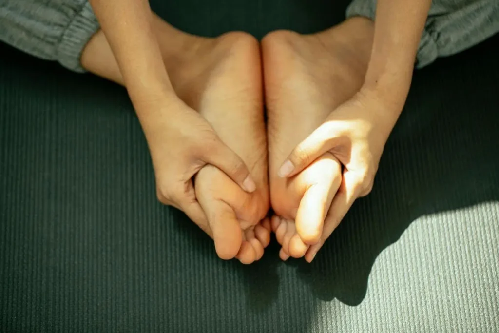 Mãos segurando os pés em posição de alongamento suave sobre tapete de yoga com luz natural representando relaxamento e recuperação do equilíbrio físico e emocional
