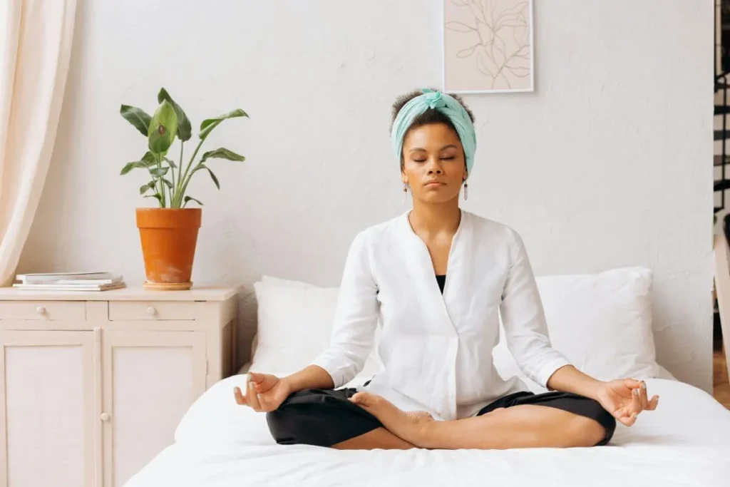 Mulher sentada na cama em posição de meditação com olhos fechados e expressão serena em quarto acolhedor com planta ao fundo