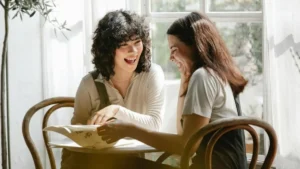 Duas mulheres sentadas à mesa rindo juntas com expressão de alegria genuína em ambiente acolhedor com luz natural
