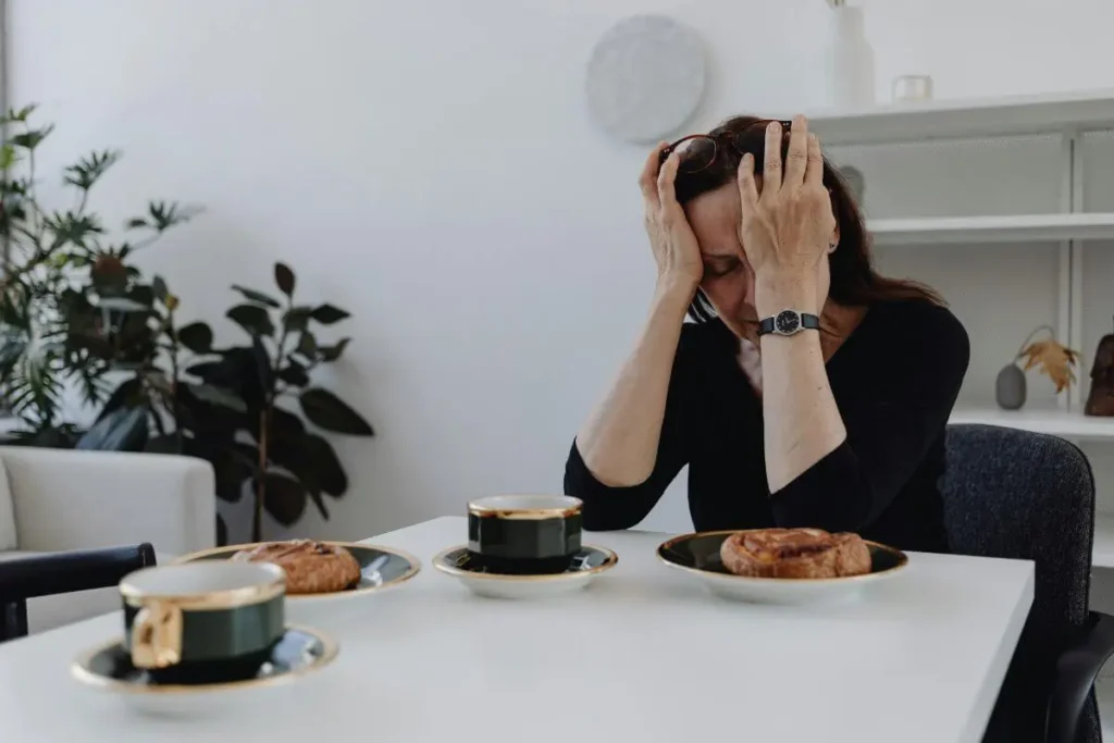 Mulher sentada à mesa com cabeça entre as mãos e expressão de tensão e estresse emocional em ambiente doméstico acolhedor