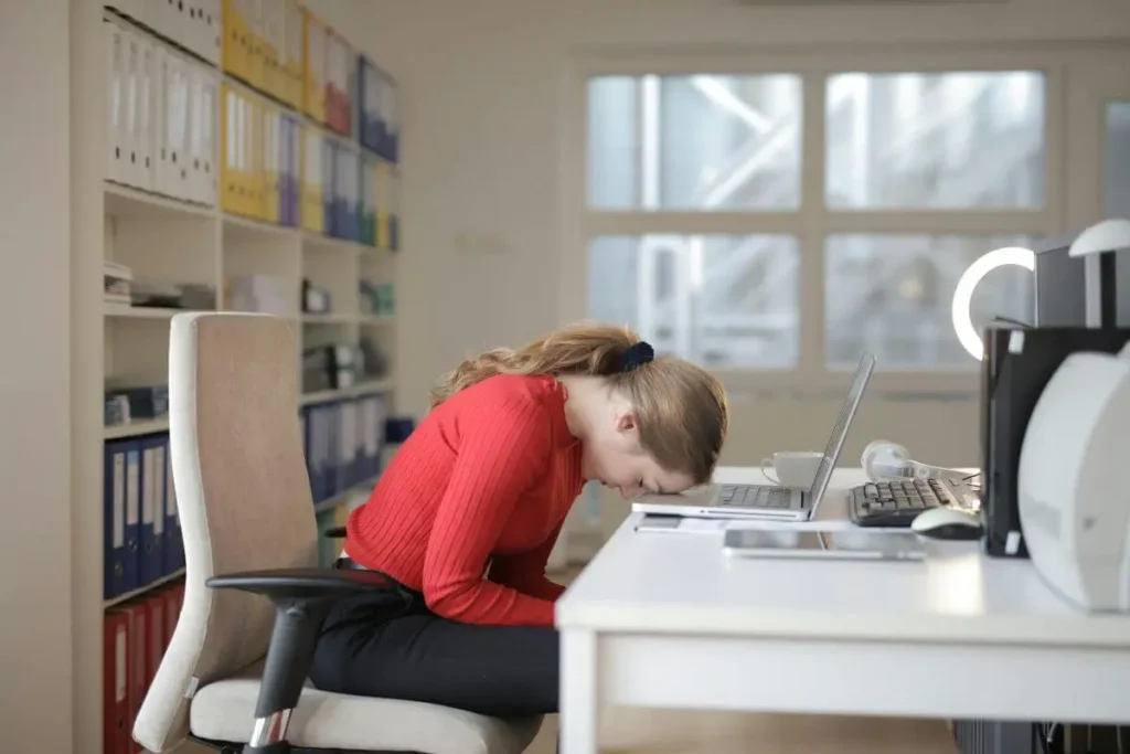 Mulher sentada em frente ao computador com postura curvada e expressão de cansaço, transmitindo sobrecarga mental e fadiga no ambiente de trabalho.