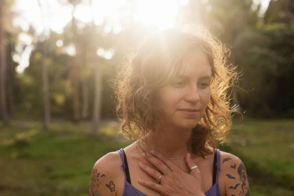 Mulher com a mão no peito em gesto de autocompaixão, expressão serena e luz natural suave ao ar livre.