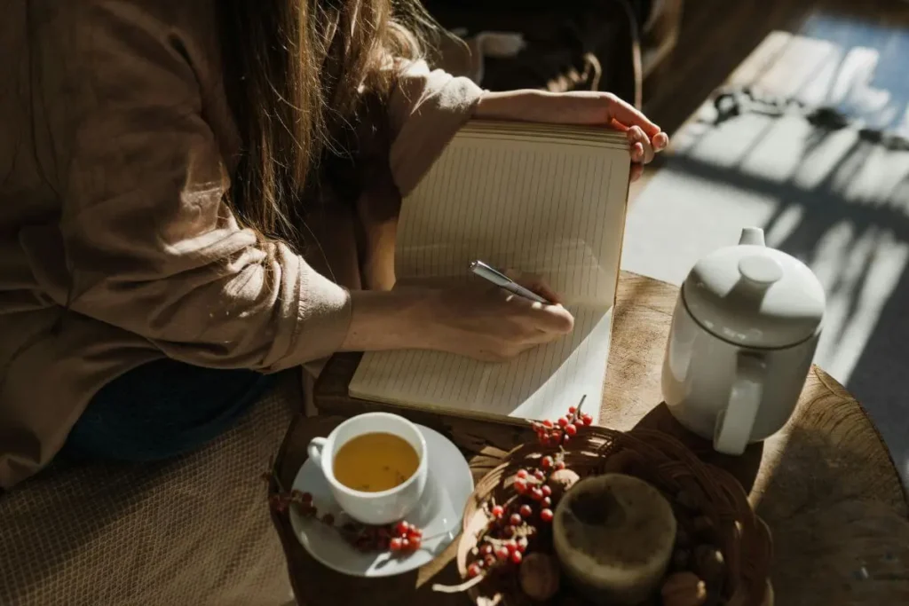 Mulher escrevendo em um caderno ao lado de uma xícara de chá, em ambiente aconchegante com luz suave.