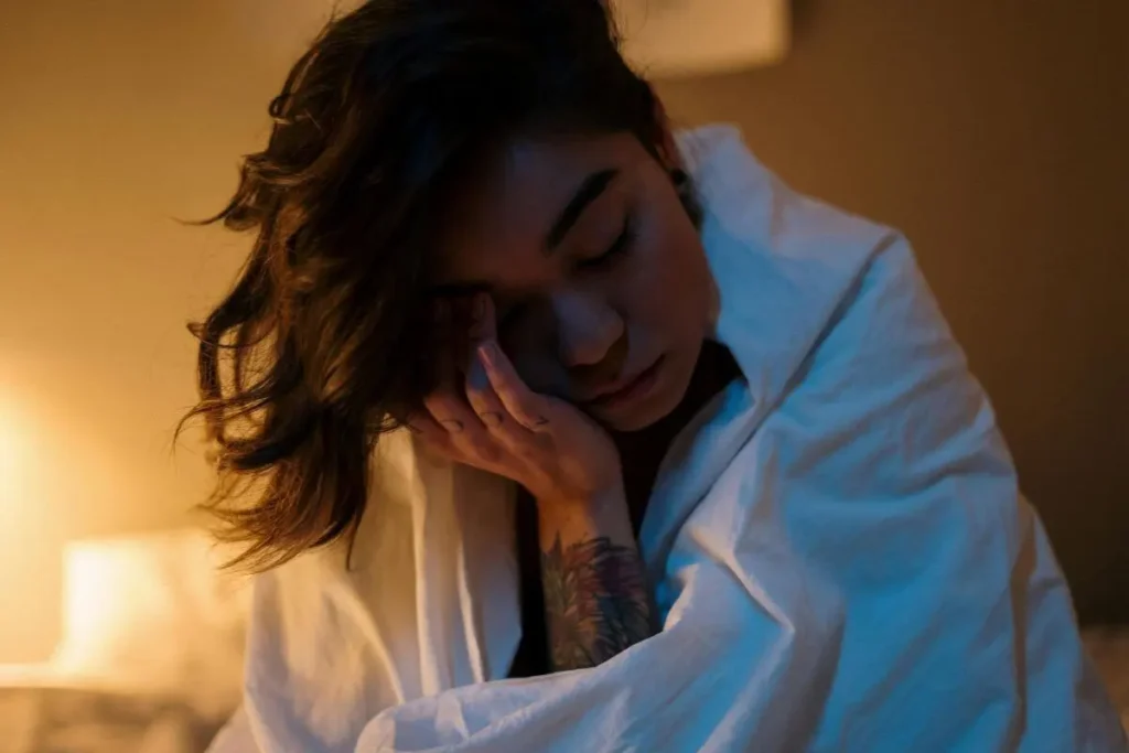 Mulher com os olhos fechados e mão apoiada no rosto, em ambiente com luz quente, expressando cansaço e introspecção.