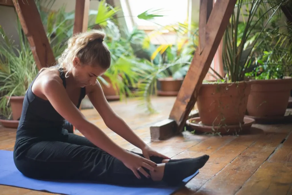 Mulher fazendo alongamento sobre tapete de yoga em ambiente doméstico com plantas ao fundo e luz natural suave.