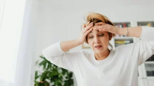 Mulher com mãos na testa e expressão de tensão e sobrecarga emocional em ambiente doméstico acolhedor com luz natural