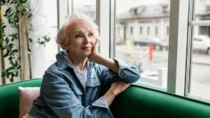 Mulher madura sentada próxima à janela, com olhar distante e expressão serena de introspecção.