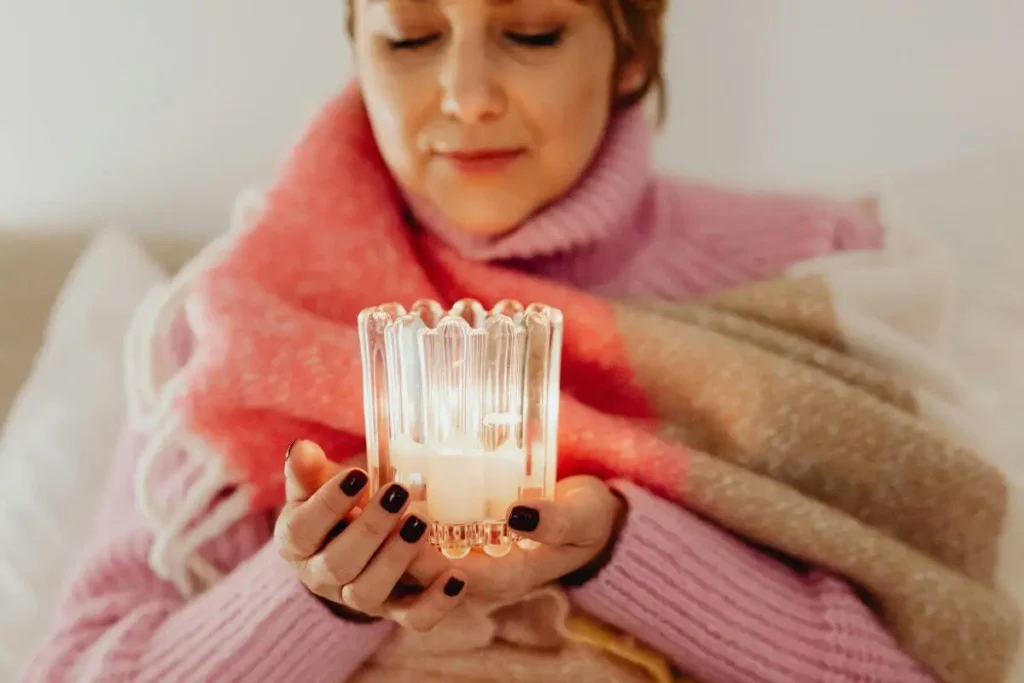 Mulher enrolada em manta segurando uma vela acesa nas mãos em ambiente acolhedor.
