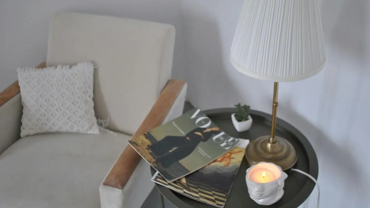 Canto aconchegante de sala com poltrona, abajur, vela acesa e livros sobre a mesa, transmitindo calma e acolhimento.