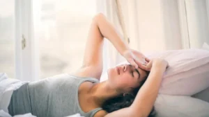 Mulher deitada na cama com mão na testa e expressão de desconforto representando ansiedade ao acordar com luz suave da manhã
