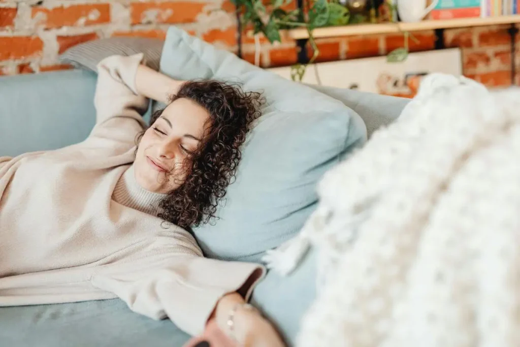Mulher deitada no sofá com olhos fechados e sorriso sereno em momento de descanso consciente em ambiente acolhedor