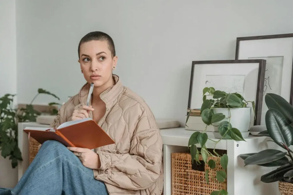 Mulher sentada escrevendo em um caderno, com expressão concentrada e reflexiva, em ambiente acolhedor com plantas ao fundo.