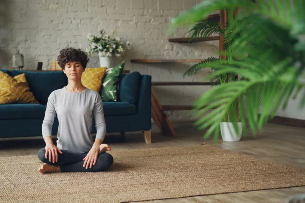 Mulher sentada em posição de meditação com os olhos fechados, praticando respiração consciente em ambiente doméstico calmo.