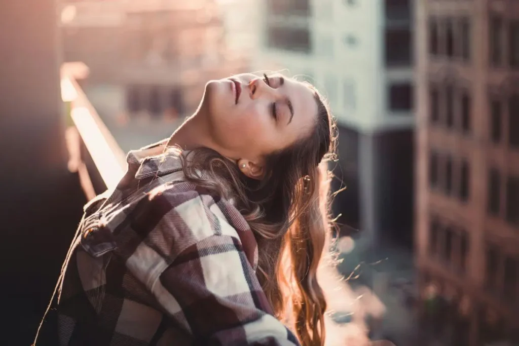mulher de olhos fechados com expressão serena absorvendo a luz do sol em momento de pausa e respiração consciente
