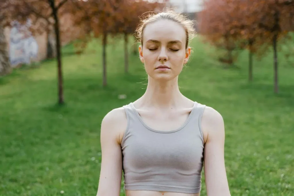 Mulher com olhos fechados meditando ao ar livre em ambiente verde, transmitindo serenidade, presença e reconexão interior.