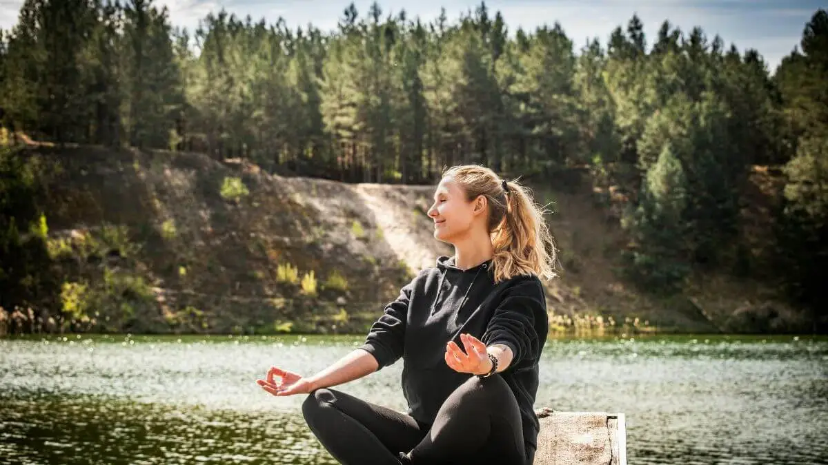Mulher meditando na natureza com olhos fechados e expressão serena, sentada à beira de um lago