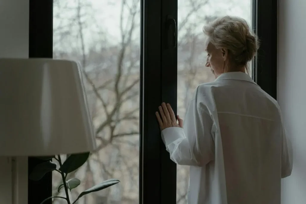 Mulher olhando pela janela em ambiente doméstico, com expressão distante e introspectiva, transmitindo desconexão emocional e reflexão silenciosa.