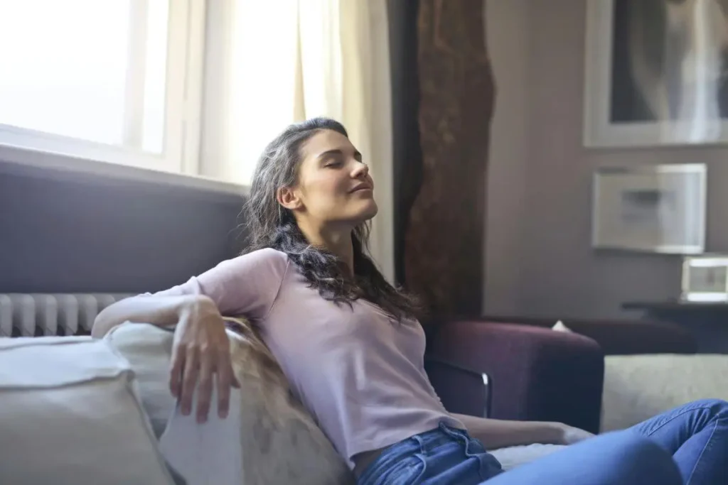Mulher recostada no sofá com olhos fechados e expressão serena, em momento de pausa consciente com luz natural