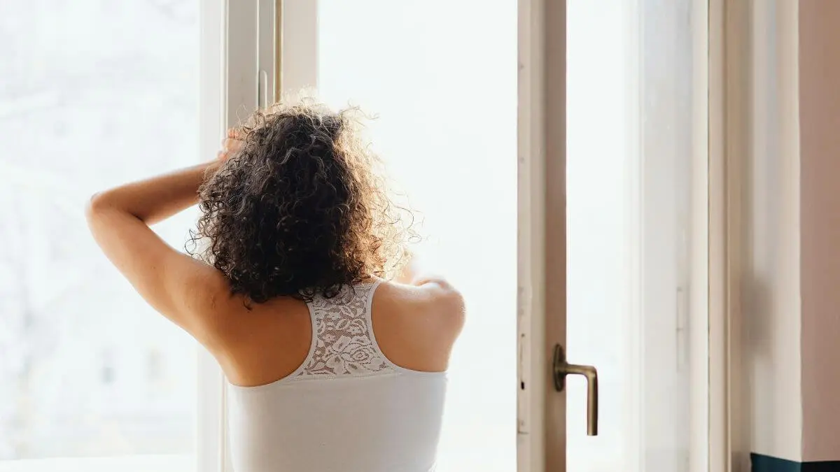 Mulher de cabelos cacheados de costas para a janela com luz natural suave representando rotina matinal para saúde mental