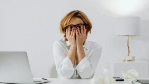 Mulher sentada à mesa de trabalho cobrindo o rosto com as mãos, demonstrando cansaço mental, estresse e sobrecarga emocional.