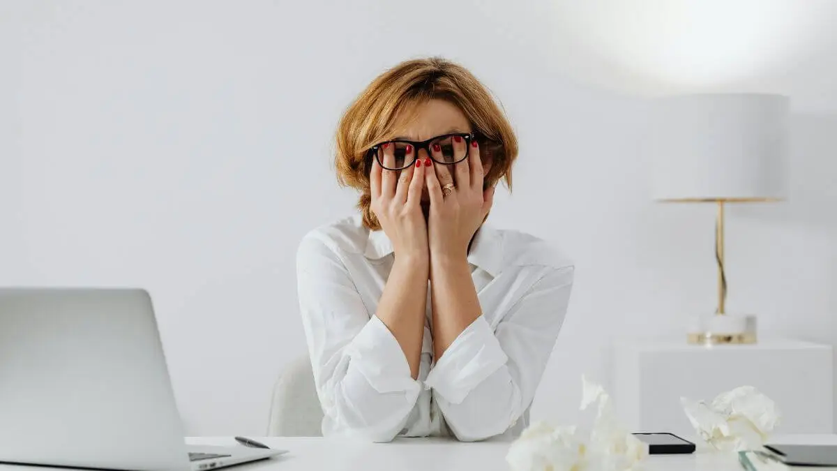 Mulher sentada à mesa de trabalho cobrindo o rosto com as mãos, demonstrando cansaço mental, estresse e sobrecarga emocional.