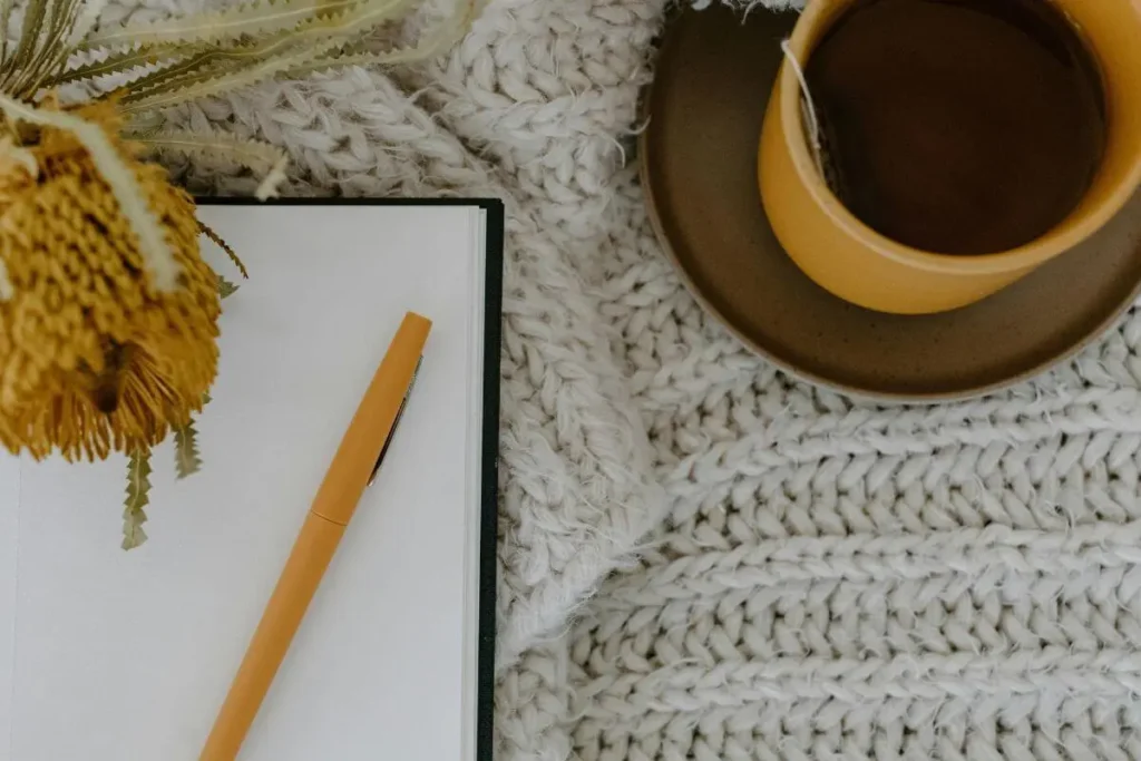 Caderno aberto com caneta ao lado de uma xícara de chá, em composição aconchegante com manta de tricô e flores secas.