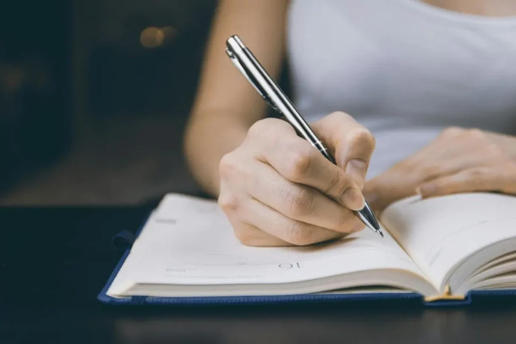 Mão de mulher escrevendo com caneta em caderno aberto, em close, representando reflexão, organização de pensamentos e escrita pessoal.